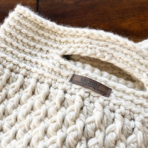 May include: A close-up of a cream-colored crocheted tote bag with a brown leather tag that reads "RUBY WEBB'S CONTEMPORARY CROCHET PATTERNS".