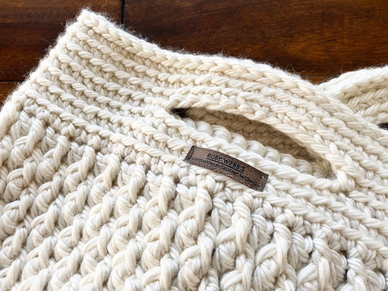 May include: A close-up of a cream-colored crocheted tote bag with a brown leather tag that reads "RUBY WEBB'S CONTEMPORARY CROCHET PATTERNS".