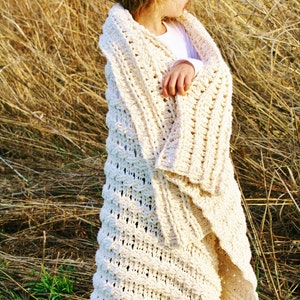 May include: A young girl wearing a white crocheted blanket in a field of tall, dry grass.