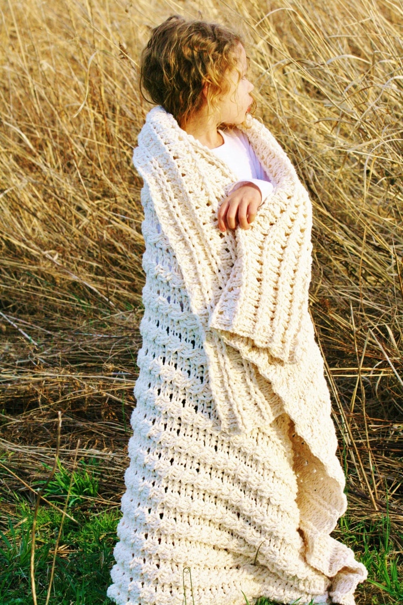 May include: A young girl wearing a white crocheted blanket in a field of tall, dry grass.