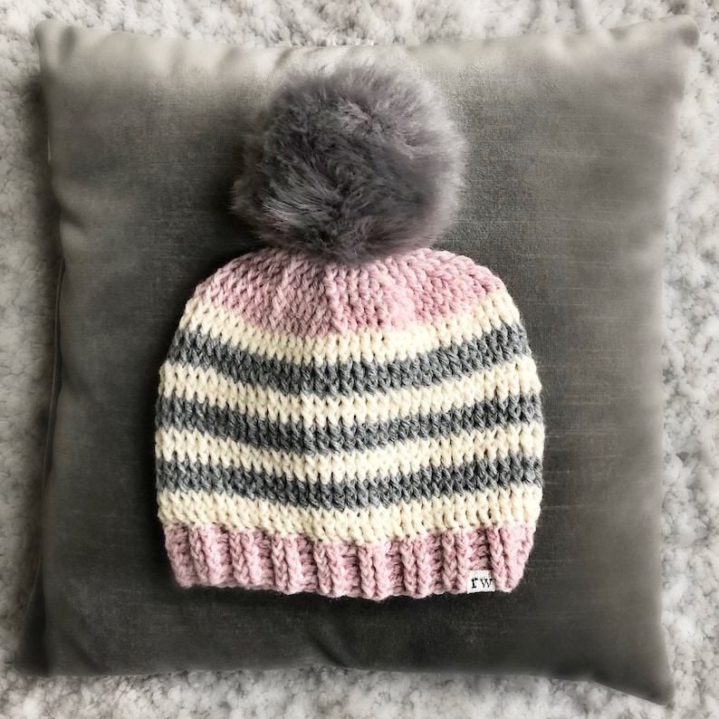 May include: A striped crocheted beanie with a pink, white, and gray color scheme. The beanie has a fluffy gray pom pom on top and a small "rw" tag on the side.