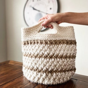 May include: A crocheted storage basket with a brown and white striped pattern and two handles. The basket is made with a thick, textured yarn.