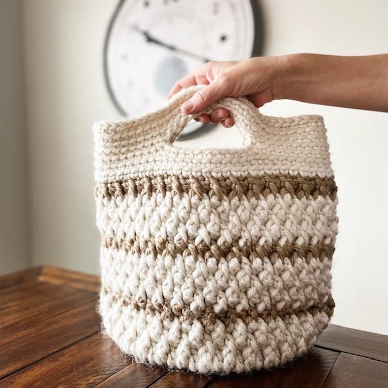 May include: A crocheted storage basket with a brown and white striped pattern and two handles. The basket is made with a thick, textured yarn.