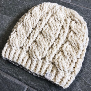 May include: A white crocheted beanie with a woven pattern. The beanie has a small black tag with the letters "I W" on it.