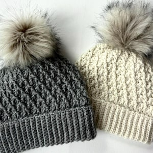 Może przedstawiać: Dwa dziane czapki z pomponami z imitacji futra. Jedna czapka jest szara, a druga kremowa.