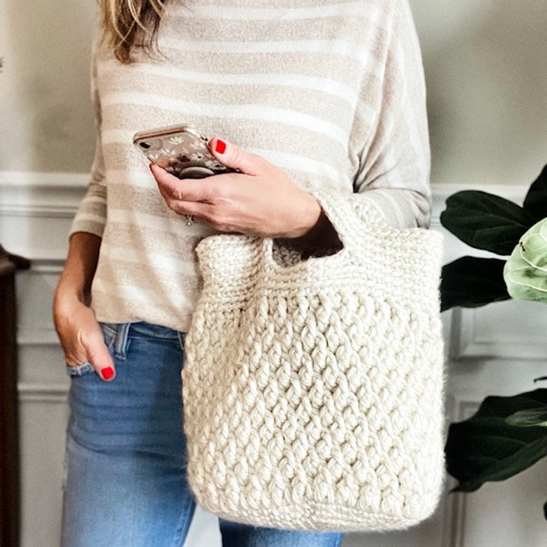May include: A white crocheted tote bag with a textured pattern. The bag has a wide, flat base and a single handle.