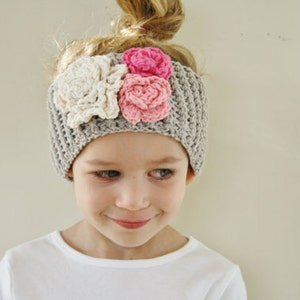 May include: A gray crocheted headband adorned with three fabric flowers in cream and pink. The headband is worn by a young person with blonde hair pulled up in a bun. The person is wearing a white shirt.