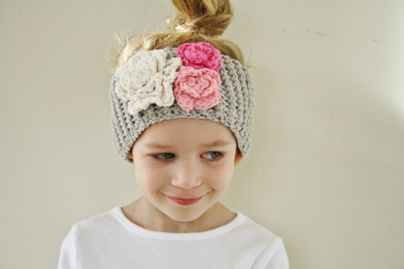 May include: A gray crocheted headband adorned with three fabric flowers in cream and pink. The headband is worn by a young person with blonde hair pulled up in a bun. The person is wearing a white shirt.