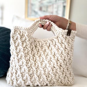 May include: Cream-colored, hand-crocheted tote bag with a textured, woven pattern. The bag features a rectangular shape with a handle and a small, brown leather tag. The bag is held by a person.