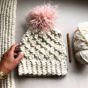 Pode incluir: Um gorro de crochê branco com um pompom rosa fofo. O gorro é feito com um ponto texturizado e tem um design simples e clássico.