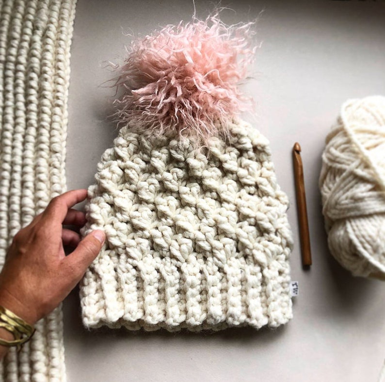 May include: A white crocheted beanie with a fluffy pink pom pom. The beanie is made with a textured stitch and has a simple, classic design.