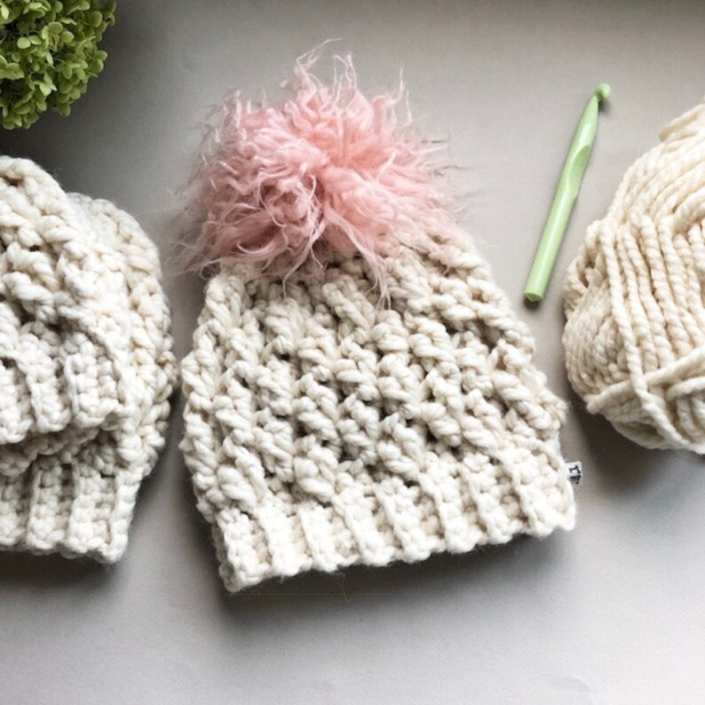 May include: Two crocheted hats with a fluffy pink pom pom on one. The hats are made with a chunky yarn and have a textured stitch pattern. A skein of yarn and a crochet hook are also in the image.