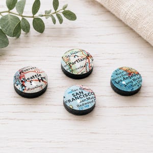 May include: Four circular magnets featuring map segments of West Coast cities including Seattle, Portland, San Francisco, and Los Angeles. Each magnet has a smooth, rounded top surface displaying printed cartographic details, city names, and road lines, set against a dark, solid base.