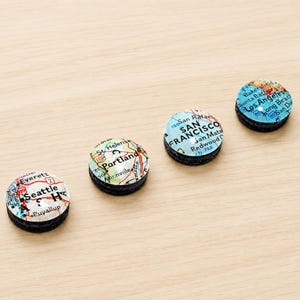 May include: Four circular magnets featuring map segments of Seattle, Portland, San Francisco, and Los Angeles. Each magnet has a clear domed top over the printed map detail and a black base, displaying city names and surrounding road networks.