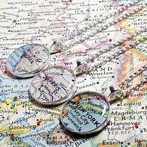 May include: Silver-toned necklaces with oval pendants featuring map designs of cities like Paris, New York, and Lima. The pendants are set against a backdrop of a detailed world map.
