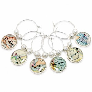 May include: A set of silver hoop wine glass charms featuring circular map inserts of various US cities including Cleveland, Las Vegas, Detroit, Boston, Omaha, and Seattle. Each charm is accented with small, textured silver-toned beads.