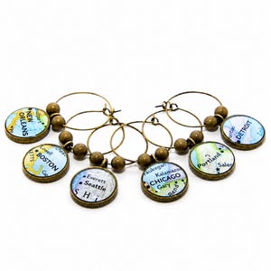 May include: A set of five circular wine glass charms featuring antique bronze metal frames and beads. Each charm displays a different map section with city names including New Orleans, Boston, Seattle, Chicago, Portland, and Detroit printed in black.