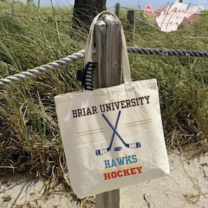 Könnte beinhalten: Eine cremefarbene Canvas-Tragetasche mit der Aufschrift "BRIAR UNIVERSITY" in Marineblau und "HAWKS Off Campus HOCKEY" in Blau und Rot. Die Tasche zeigt gekreuzte Hockeyschläger und einen Puck. Die Tasche hängt an einem Holzpfosten.