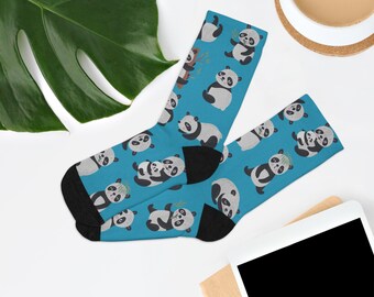 Op de afbeelding: Een paar blauwe sokken met een herhaald patroon van zwart-witte panda's. De sokken hebben zwarte hielen en tenen. De achtergrond is wit met een groot groen blad, een kopje koffie en een tablet.