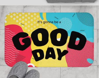 Puede incluir: Una alfombra de baño rectangular con un diseño colorido. La alfombra presenta las palabras "GOOD DAY" en letras negras grandes. El fondo tiene una mezcla de formas rojas, amarillas y azules, con la frase "It's gonna be a" encima del texto principal.