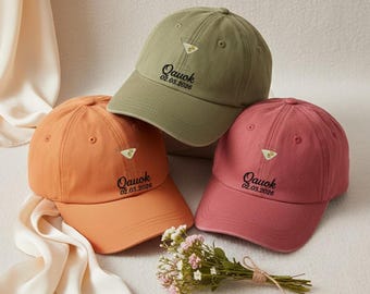 May include: Three baseball caps in orange, olive green, and rose. Each cap has a martini glass graphic and the text "Qauok 02.03.2026". A small bouquet of flowers is near the caps.