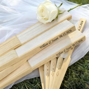May include: Several personalized wooden fans with white paper inserts, engraved with "William & Susan 06.12.2026". A white rose rests on top of the fans. The fans are arranged on a white cloth, with a green background.