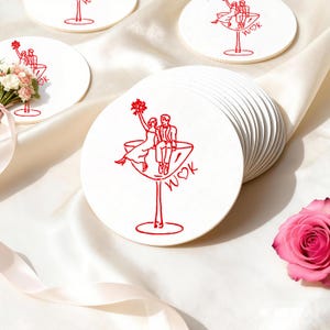 Puede incluir: Posavasos redondos blancos con una ilustración roja de una pareja en una copa de cóctel. El diseño incluye las iniciales "W♡K". Se ve una pila de posavasos, junto con una rosa rosa y un ramo de flores.