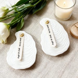 May include: Two white, seashell-shaped ring dishes with a pearl accent and a small wooden tag. The tag is engraved with the text "Asqit & Exih 09.05.2026". A lit candle, a ring box, and white flowers are in the background.