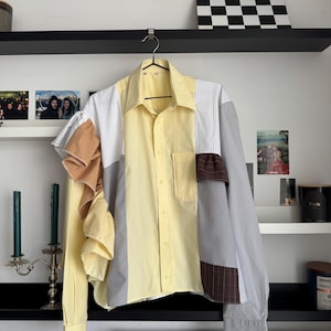 May include: A patchwork button-down shirt with a yellow, white, gray, and brown color scheme. The shirt features a unique design with various fabric patterns and textures, including ruffles on one side.