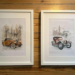 May include: Two framed cross-stitch artworks. One features a vintage car in a city scene with a clock tower. The other depicts a classic car in front of a landmark building. Both are in white frames, displayed on a wooden surface.