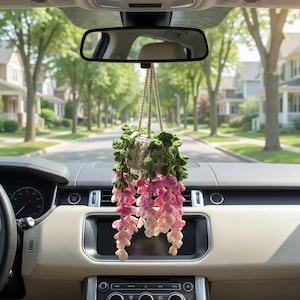 May include: A handmade crochet hanging plant in a car. The plant features a beige pot, green leaves, and pink and peach-colored cascading flowers. The plant hangs from the rearview mirror, adding a touch of nature to the car's interior.
