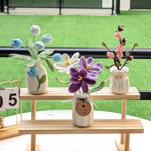 May include: Hand-crocheted flower arrangements in small vases, displayed on a wooden shelf. The flowers include blue, white, and purple blooms. One vase has a tag that reads "YOU ARE LOVED". The background is a green lawn.