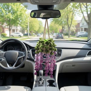 May include: A crocheted hanging plant ornament suspended from the rearview mirror of a vehicle. The piece features a small beige pot containing green foliage and long, cascading strands of purple and lavender flowers made of yarn.