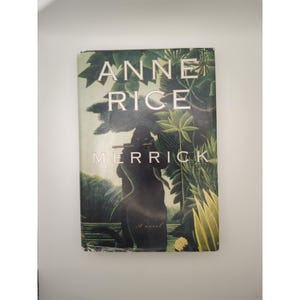 May include: A hardcover book titled "ANNE RICE MERRICK" with a jungle-themed cover. The cover features a dark silhouette figure playing a flute, surrounded by lush green foliage. The author's name and book title are in white text.