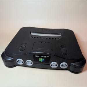 Peut inclure: Une console de jeux vidéo Nintendo 64 noire dotée d'un port cartouche frontal, de quatre ports manette, d'un bouton d'alimentation, d'un bouton de réinitialisation et d'un cache pour l'extension mémoire. Le logo NINTENDO 64 est visible.
