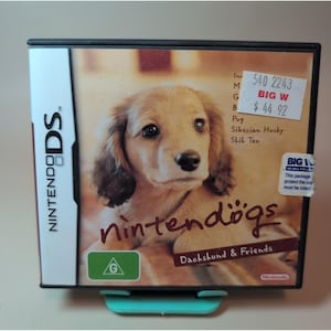 May include: A Nintendo DS game case for Nintendogs: Dachshund & Friends featuring a light brown puppy on the cover. The case includes a white spine with the Nintendo DS logo, a green G rating icon, and two retail stickers on the front surface.