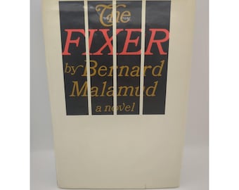 以下が含まれることがあります： バーナード・マラマッド著「The Fixer」の表紙。タイトルは赤色で、著者の名前と「a novel」は金色です。背景はクリーム色で、黒い縦棒があります。