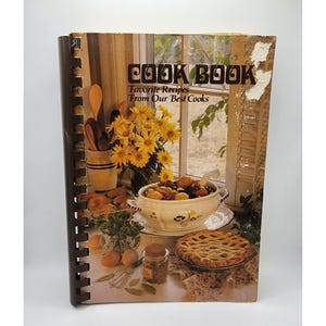 Puede incluir: Un libro de cocina vintage con encuadernación en espiral. La portada muestra las palabras "COOK BOOK" y "Favorite Recipes From Our Best Cooks". La imagen de la portada muestra una naturaleza muerta de comida, flores y una ventana.