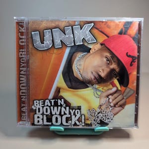 May include: A CD case featuring the artist UNK. The album cover has a portrait of the artist wearing a red cap and a silver chain. The album title "Beat N Down Yo Block!" is displayed in bold text.