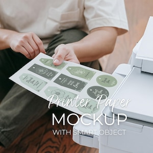 Peut inclure: Une personne tient une feuille d'étiquettes imprimées avec des motifs floraux et du texte, à côté d'une imprimante blanche. Les étiquettes sont dans des tons de vert et de gris, avec des phrases comme "Merci" et "Fait avec amour". L'image comprend le texte "Printer Paper Mockup with Smart Object".