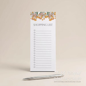 May include: A white notepad with a floral design at the top, labeled "SHOPPING LIST." The notepad is 3.5 x 8.5 inches. A silver pen rests in front of the notepad. The notepad has a series of circles and lines for a shopping list.