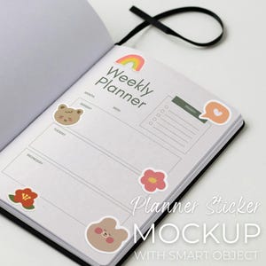 May include: Open black notebook with a weekly planner layout. The page is decorated with colorful stickers, including a rainbow, flowers, and bear-shaped designs. Text on the page reads "Weekly Planner" and "Planner Sticker MOCKUP WITH SMART OBJECT."