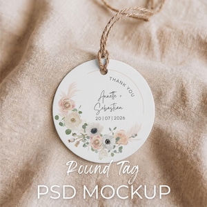 May include: A round white tag with a floral design and the text "Thank You Annette + Sebastian 20|07|2026". The tag is suspended by a natural twine string. The words "Round Tag PSD MOCKUP" are at the bottom.