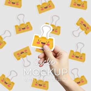 May include: A hand holding a yellow sticker with a smiling face and a binder clip design. The sticker is part of a mockup with the text "Sticker MOCKUP WITH SMART OBJECT" in white. Other similar stickers are scattered in the background.