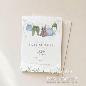 Puede incluir: Maqueta de tarjeta de invitación para baby shower. La tarjeta presenta ilustraciones de acuarela de ropa de bebé colgada en un tendedero. El texto dice "Join us for a Baby Shower Celebrating AML" con detalles de fecha y hora. Tarjeta 5x7.