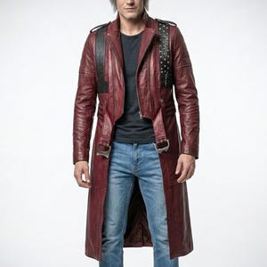 May include: A burgundy leather trench coat with a black shoulder strap and silver buckles. The coat has a zipper closure and a long, flowing design. The model is wearing a black t-shirt and blue jeans.