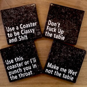 May include: Set of four square coasters with a dark speckled design. Each coaster has a different white text message: "Use a Coaster to be Classy and Shit", "Don't Fuck Up the table", "Use this coaster or I'll punch you in the throat", and "Make me Wet not the table".