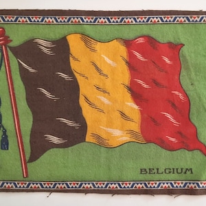 May include: A vintage, rectangular, fabric patch with a brown, yellow, and red Belgian flag on a green background. The flag is on a pole with tassels. The patch has a brown border with a decorative pattern and the word "BELGIUM" printed in black at the bottom.
