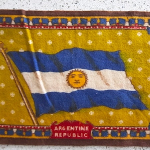 May include: A vintage fabric patch with a depiction of the Argentine flag. The flag is blue and white with a yellow sun in the center. The text "ARGENTINE REPUBLIC" is printed below the flag.