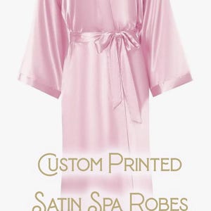 May include: A light pink satin spa robe with a matching tie belt. The robe has wide sleeves and a V-neck. The text "The Sisterhood" is printed on the upper back. The words "Custom Printed Satin Spa Robes" are printed at the bottom.